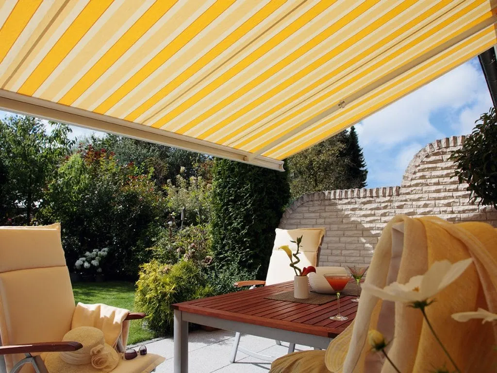 Eine sonnige Gartenterrasse mit einer ausgedehnten gelb-weiß gestreiften Markise, bequemen Stühlen und einem Tisch mit einer Blumenvase.