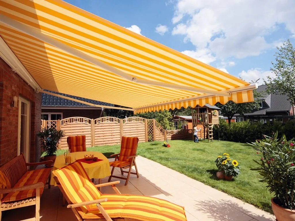 Eine sonnige Terrasse im Hinterhof, beschattet von einer großen gestreiften Primasol Markise-Markise-Markise mit Gartenmöbeln und Topfpflanzen.