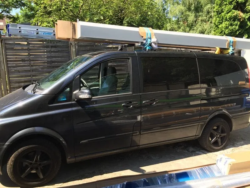 Ein schwarzer Lieferwagen mit Baumaterialien auf dem Dachträger, der neben Baumaterialien geparkt ist.