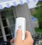 Hand hält eine Fernbedienung, die auf ein Haus mit Markise gerichtet ist.