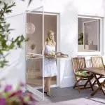 Eine Frau mit einem Tablett betritt eine sonnige Terrasse, die mit Insektenschutz ausgestattet und für eine Mahlzeit vorbereitet ist.