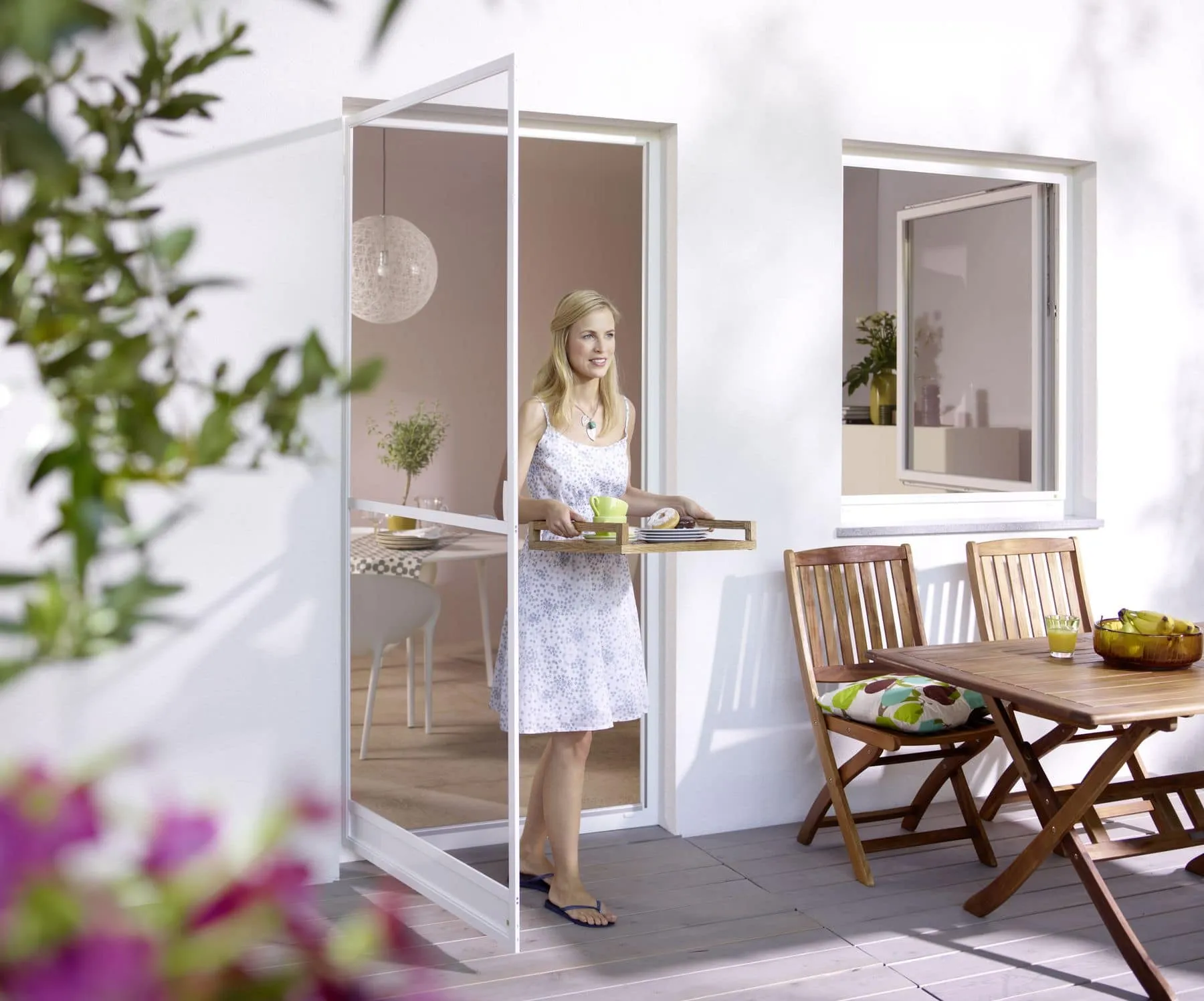 Eine Frau mit einem Tablett betritt eine sonnige Terrasse, auf der mit Insektenschutz eine Mahlzeit serviert wird.