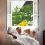Ein Paar ruht sich im Bett aus. Das offene, durch Insektenschutz geschützte Fenster bietet einen Blick auf einen Garten und die Außenansicht eines Gebäudes.