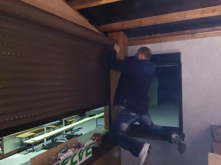 Eine Person tritt durch ein Fenster in einen dunklen Raum mit teilweise hochgeklappten Fensterläden.