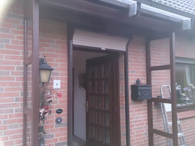 Vordereingang eines Hauses mit einer Holztür und einer Backsteinfassade sowie einer Außenleuchte und einem Briefkasten.