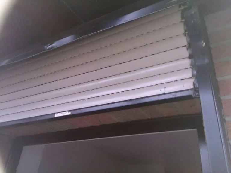 Ein teilweise heruntergelassener beigefarbener Fensterladen mit einigen Gebrauchsspuren und sichtbarem Schmutz, installiert über einem Fensterrahmen in einer Backsteinmauer.