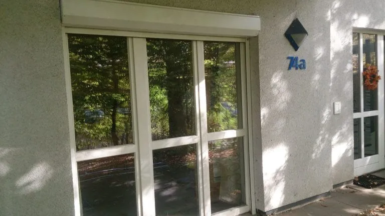 Ein Glastüreingang eines Gebäudes mit der Nummer 7a daneben, umgeben von grünem Laub.