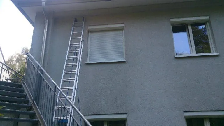 Metallleiter lehnt an einem zweistöckigen Gebäude mit geschlossenen Fensterläden.