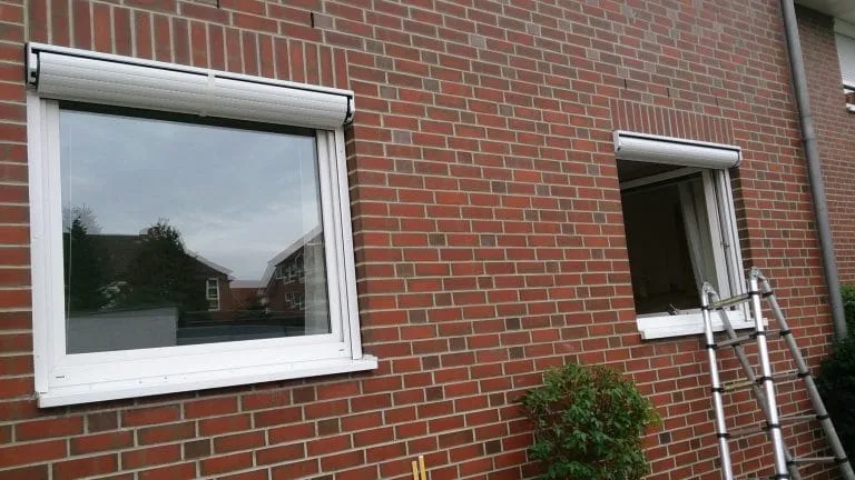 Zwei Fenster in einer Backsteinmauer; eines ist geschlossen, das andere offen und draußen steht eine Leiter.