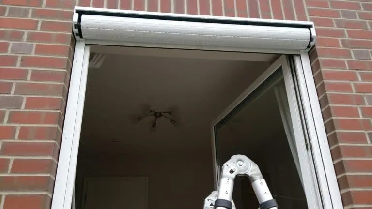 Ein offenes Fenster mit teilweise heruntergelassenem Rollladen und einem Deckenventilator, der sich im Glas spiegelt.