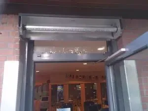 Luftschleiergerät, das über einem Eingang zu einem Gewerbebetrieb installiert wird und mit sichtbaren, selbstklebenden Vogelabwehrspikes ausgestattet ist, um das Sitzen von Vögeln zu verhindern.