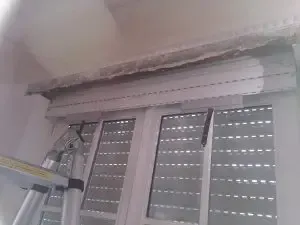 Beschädigte Decke über einem Fenster mit teilweise freiliegendem Sturz und einer Leiter in der Nähe des Fensters.