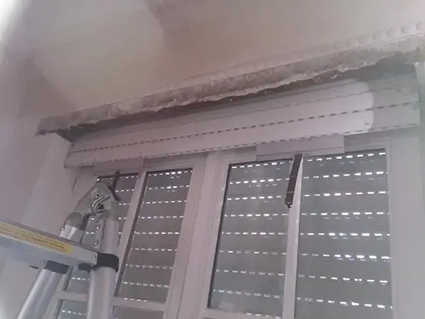 Beschädigte Decke über einem Fenster mit teilweise freiliegendem Sturz und einer Leiter in der Nähe des Fensters.